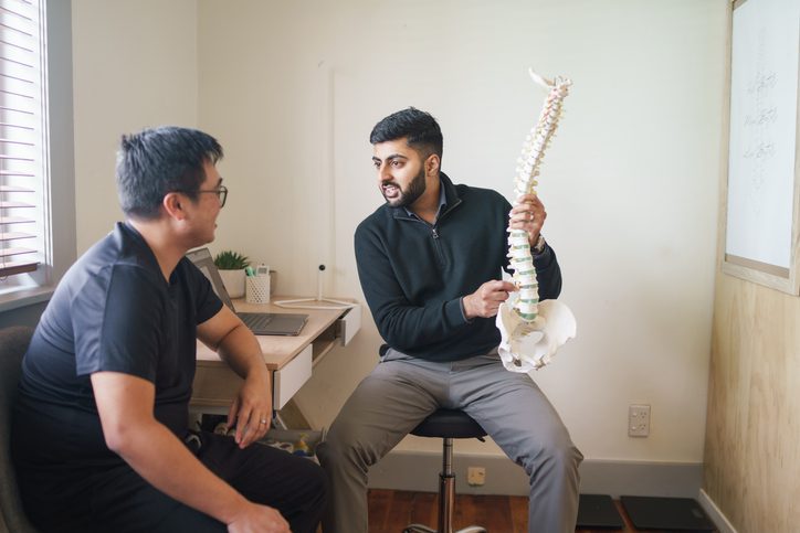 Chiropractor explaining spinal anatomy to a patient in a medical consultation room with a spine model.